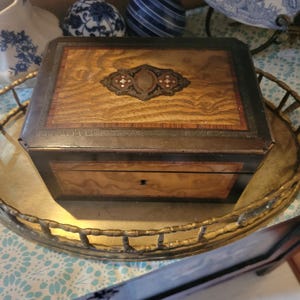 May include: A rectangular wooden box featuring a dark brown frame with a Greek key pattern border. The lid displays a light brown wood grain center panel with an ornate, dark, inlaid decorative plaque. The front panel has a keyhole.