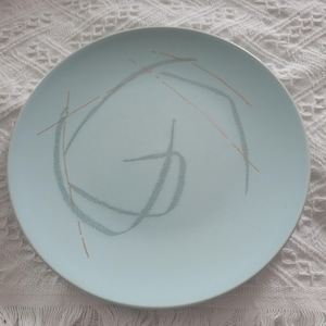 May include: A light blue ceramic plate with a mid-century modern design. The plate features a gray and gold abstract pattern. The plate is round and has a smooth surface. This plate is perfect for serving food.