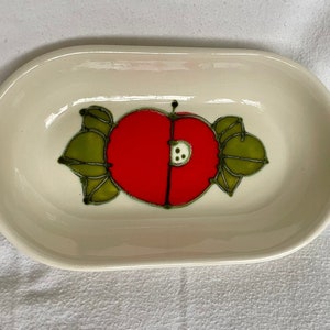 May include: Cream-colored ceramic serving dish with an oval shape. The dish features a red apple design with green leaves and black outlines. The apple design is centered on the dish.