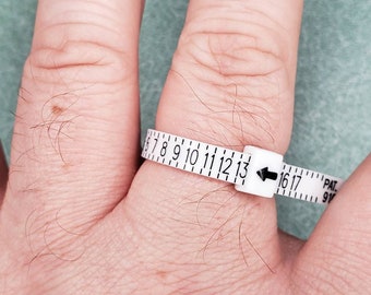 May include: A white plastic ring sizer with black numbers and an arrow pointing to the size 13. The ring sizer is on a person's finger.