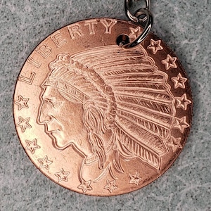 May include: A copper-colored round keychain pendant featuring a detailed profile of a Native American in a headdress, with the word "LIBERTY" above. The pendant is ringed with stars and attached to a silver-colored chain.