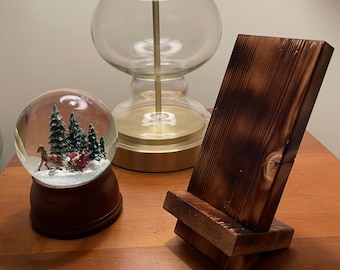 May include: A wooden phone stand with a dark brown finish. The stand is made of two pieces of wood, one stacked on top of the other. The stand is designed to hold a phone upright.