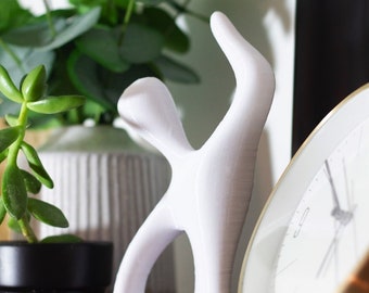 May include: A white abstract sculpture of a person with arms raised, standing on a wooden surface. A gold-rimmed clock and potted plants are in the background. The sculpture is approximately 6 inches tall.