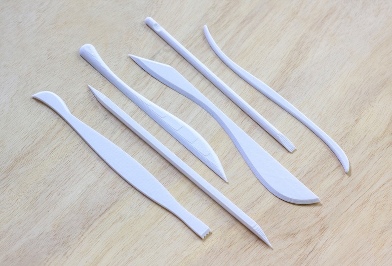 May include: Set of seven white plastic sculpting tools with various shapes and sizes. The tools are laid out on a light wood surface.