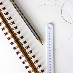 May include: A white spiral bound notebook with a pencil and a white plastic ruler with holes on it. The notebook is open to a blank page with a few circles drawn on it.
