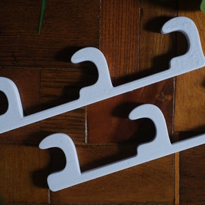 Może przedstawiać: Dwa białe plastikowe haczyki z trzema zębami każdy. Haczyki są przeznaczone do zawieszania przedmiotów na ścianie lub innej powierzchni.