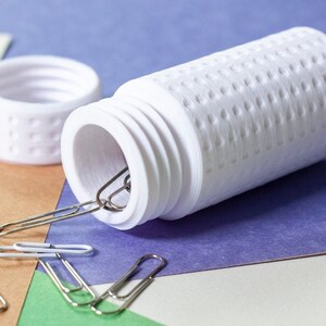 Op de afbeelding: Een witte plastic container met een getextureerd oppervlak, open en met zilveren paperclips erin. De container heeft een deksel.