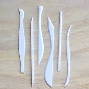 May include: Set of seven white plastic sculpting tools for crafting and modeling clay. The tools have different shapes and sizes, including a curved blade, a pointed tool, and a flat tool.