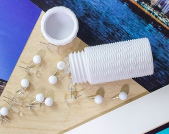 May include: A white plastic container with a ribbed design, filled with white push pins. The container is open and some push pins are scattered on a wooden surface.