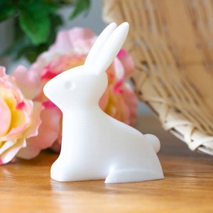 Könnte beinhalten: Eine weiße Keramik-Hasenfigur mit schlichtem Design. Der Hase sitzt auf einer Holzoberfläche mit rosa Rosen im Hintergrund.