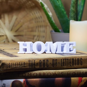 Może przedstawiać: Białe drewniane litery układające się w słowo "HOME" są ułożone na stosie trzech starych książek. Książki mają brązowe okładki z tekstem na grzbietach. Na tle znajduje się doniczka z aloesem.