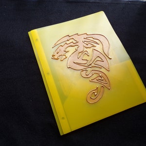 May include: A yellow plastic folder with a copper-colored dragon design. The folder has three holes for a binder.
