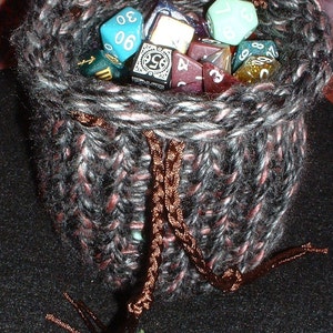 Pode incluir: Uma bolsa de cordão de malha preta e rosa cheia de dados coloridos. Os dados são de várias formas e tamanhos, com números e símbolos em suas faces.