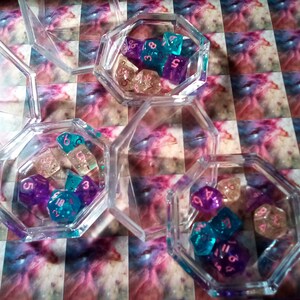 May include: Three clear octagonal containers filled with colorful dice. The dice are translucent and have pink numbers. The dice are in shades of purple, blue, and white.