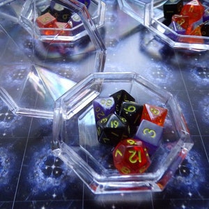 May include: A clear plastic octagonal box filled with colorful dice. The dice are red, black, purple, and white with black numbers. The box is sitting on a blue and white patterned surface.