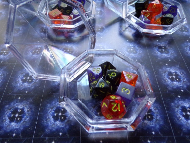 May include: A clear plastic octagonal box filled with colorful dice. The dice are red, black, purple, and white with black numbers. The box is sitting on a blue and white patterned surface.