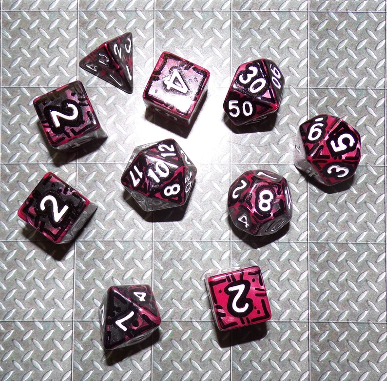May include: A collection of ten polyhedral dice, each with a unique shape and number of sides. The dice are clear with a pink and black design, and white numbers. The dice are arranged on a silver, diamond-plate surface.