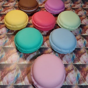 May include: Nine colorful macarons arranged in a semi-circle on a patterned surface. The macarons are in shades of pink, green, blue, yellow, brown, and light green.
