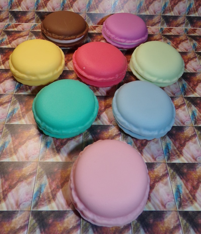 May include: Nine colorful macarons arranged in a semi-circle on a patterned surface. The macarons are in shades of pink, green, blue, yellow, brown, and light green.