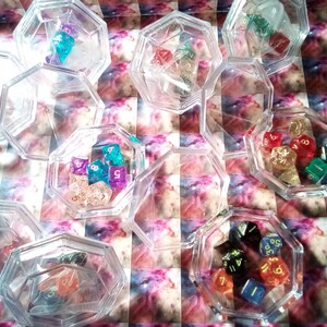 Może przedstawiać: Kolekcja przezroczystych plastikowych pojemników wypełnionych kolorowymi kostkami do gry. Kostki są różnych kształtów i rozmiarów, niektóre mają metaliczne wykończenie.