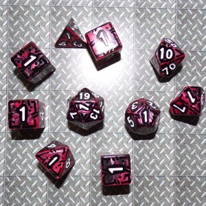 May include: A collection of ten polyhedral dice, each with a clear body and red and black markings. The dice are arranged on a silver diamond-plate surface. The numbers are white.