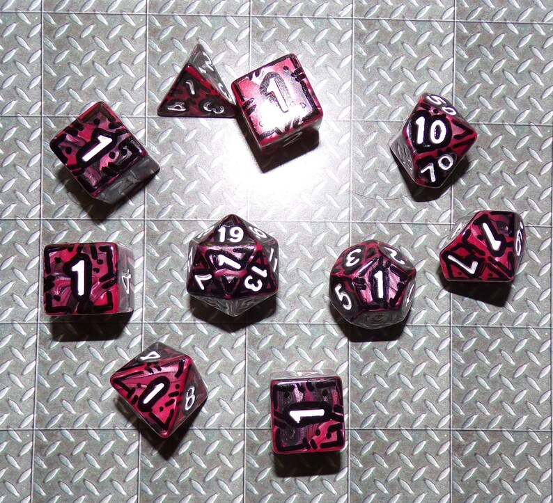 May include: A collection of ten polyhedral dice, each with a clear body and red and black markings. The dice are arranged on a silver diamond-plate surface. The numbers are white.