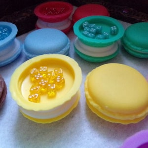 May include: Assortment of colorful macarons with various fillings, including yellow dice, blue dice, and red sprinkles. The macarons are arranged on a white surface.
