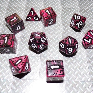 May include: A collection of ten polyhedral dice in various shapes, including a triangular prism, cubes, and a dodecahedron. The dice are transparent with a red and black design and white numbers. The dice are for tabletop games.
