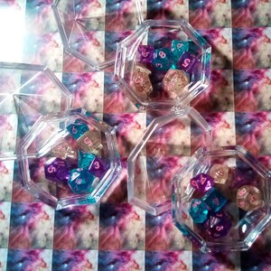 May include: A set of six clear plastic containers with lids, each containing a collection of colorful dice. The dice are a mix of purple, blue, and pink with white numbers.