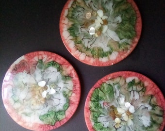 Puede incluir: Tres posavasos redondos con borde rojo y diseño abstracto blanco, verde y dorado. Cada posavasos tiene pequeños detalles blancos y dorados incrustados en la resina.
