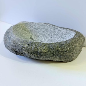 Peut inclure: Un mortier en pierre grise avec un extérieur rugueux et texturé et un intérieur lisse en forme de bol. Le mortier est de forme ovale et semble sculpté à la main. La pierre présente des nuances de gris et de vert.