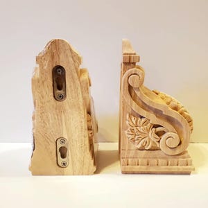 Peut inclure: Deux consoles en bois non finies, côte à côte, sur fond blanc. Les consoles sont sculptées avec des détails décoratifs, notamment des motifs floraux et des volutes. Chaque console a des trous pré-percés pour le montage.