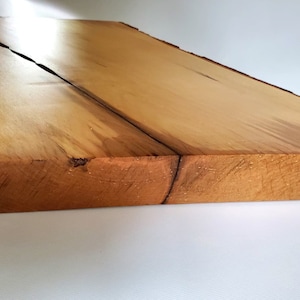 May include: A large, rectangular wooden board with a natural wood finish. The board has a live edge on one side and a smooth, polished surface. The board is made of two pieces of wood that have been joined together.