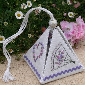 May include: A white fabric pyramid-shaped box with a purple trim and embroidered floral designs. The box has a white tassel and a white cord.