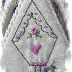 May include: White fabric with a green and gold embroidered design. The design features a heart, a flower, and a series of small dots.