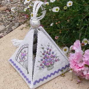 May include: A white fabric pyramid-shaped box with a white tassel and a silver-coloured top. The box is decorated with embroidered flowers and a fan design. The box is sitting on a light brown surface.