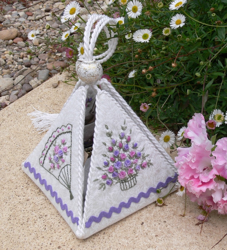 May include: A white fabric pyramid-shaped box with a white tassel and a silver-coloured top. The box is decorated with embroidered flowers and a fan design. The box is sitting on a light brown surface.