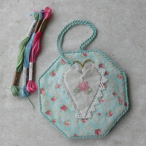 May include: A hexagonal fabric ornament featuring a light blue floral pattern with pink roses, trimmed with twisted light blue cord. A central heart-shaped patch with white lace edging displays an embroidered pink lotus flower, accompanied by three skeins of embroidery floss.