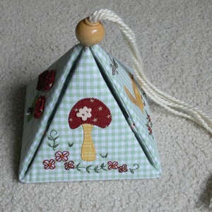 Peut inclure: Une bo&icirc;te en forme de pyramide en tissu vichy vert et blanc avec un champignon, des fleurs et des oiseaux brod&eacute;s. La bo&icirc;te a un cordon blanc avec un bouton en bois sur le dessus.