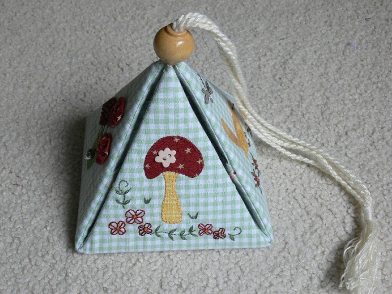 Peut inclure: Une bo&icirc;te en forme de pyramide en tissu vichy vert et blanc avec un champignon, des fleurs et des oiseaux brod&eacute;s. La bo&icirc;te a un cordon blanc avec un bouton en bois sur le dessus.