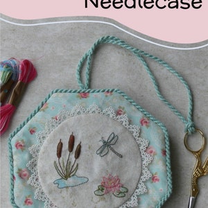 May include: A hexagonal needlecase featuring a light blue fabric with a pink rose pattern, bordered by a twisted mint green cord. The center displays an embroidered circle with cattails, a dragonfly, and a water lily, framed by white lace.