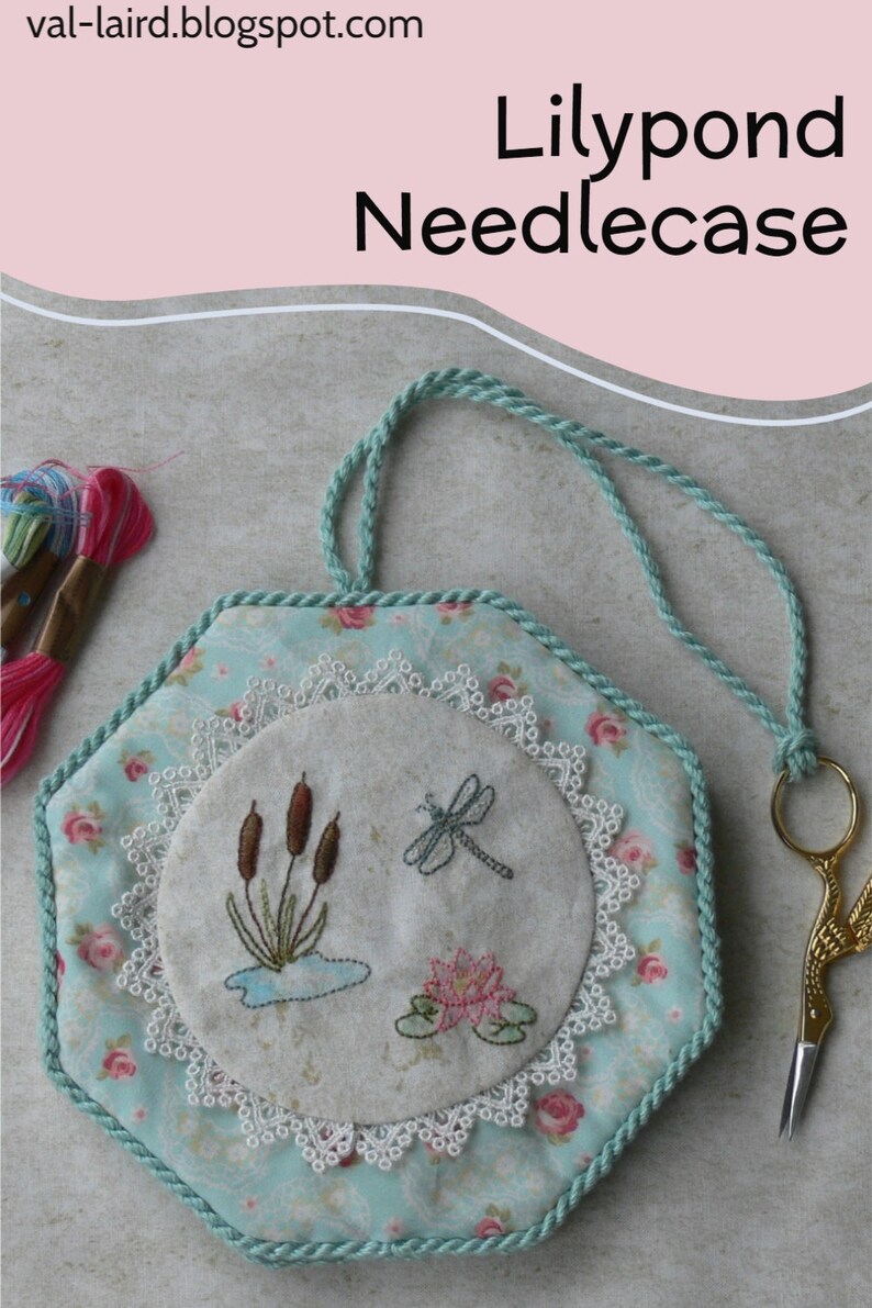 May include: A hexagonal needlecase featuring a light blue fabric with a pink rose pattern, bordered by a twisted mint green cord. The center displays an embroidered circle with cattails, a dragonfly, and a water lily, framed by white lace.