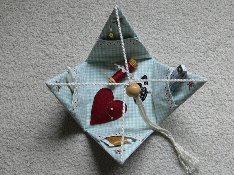 Peut inclure: Un kit de couture en tissu vichy vert et blanc avec un c&oelig;ur rouge, une bobine de fil et un coussin &agrave; aiguilles. Le kit est reli&eacute; par une ficelle blanche.