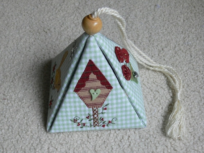 Peut inclure: Une petite bo&icirc;te en tissu de forme de maison en vichy vert et blanc avec un motif de nichoir rouge et brun sur le devant. La bo&icirc;te a un cordon blanc avec un bouton en bois sur le dessus et un pompon &agrave; l'extr&eacute;mit&eacute;.