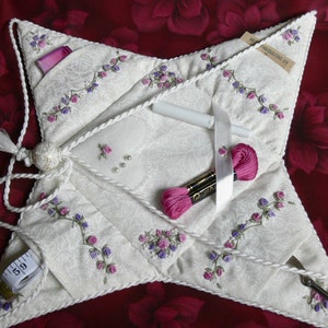 May include: A white star-shaped sewing kit with a pink and purple floral embroidery design. The kit has multiple pockets and includes a tape measure, scissors, thread, and ribbon.