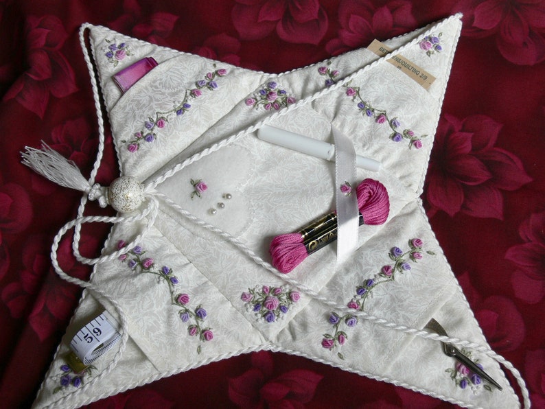 May include: A white star-shaped sewing kit with a pink and purple floral embroidery design. The kit has multiple pockets and includes a tape measure, scissors, thread, and ribbon.