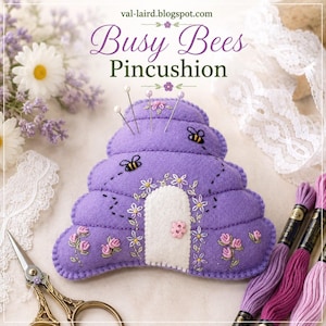 May include: A purple felt pincushion shaped like a beehive, featuring embroidered bees, flowers, and a white felt door with a pink button. The text "Busy Bees Pincushion" and "val-laird.blogspot.com" is printed above the object.