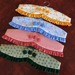 May include: Four fabric-covered hangers with ruffled edges. The hangers are in different colors and patterns, including yellow, blue, pink, and green.