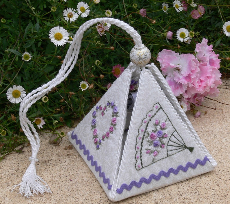 May include: A white fabric pyramid-shaped box with a purple trim and embroidered floral designs. The box has a white tassel and a white cord.