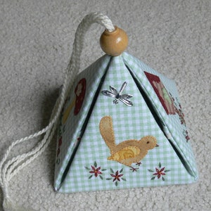 Peut inclure: Une bo&icirc;te en forme de pyramide en tissu vichy vert et blanc avec un cordon blanc et un bouton en bois. La bo&icirc;te est d&eacute;cor&eacute;e de d&eacute;tails brod&eacute;s d'oiseaux, de fleurs et d'une libellule.